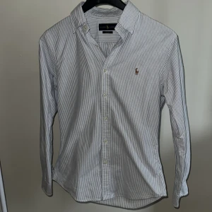skjorta från Ralph Lauren - skjorta från Ralph Lauren i blå och vit randigt mönster. Väldigt bra skick, använd enstaka gånger förra sommaren. Perfekta skjortan inför sommaren. Har även fler skjortor som inte är utlagda (kontakta för info)