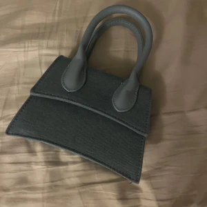 Ljusblå miniväska med axelrem - Säljer en stilren mörkgrå miniväska med både handtag och avtagbar axelrem. Väskan har en unik, snedskuren design och är tillverkad i ett tygmaterial med detaljer i syntet. Perfekt för att bära det viktigaste och ge outfiten en cool touch.