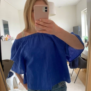 Blå offshoulder blus - Säljer en blå offshoulder blus med vida ärmar och lätt struktur i tyget. Toppen har en lös passform och är perfekt för varmare dagar. Snygg att styla med jeans eller kjol för en trendig look.