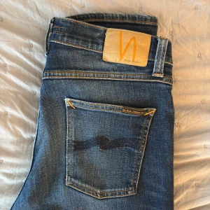 Mörkblå jeans från Nudie Jeans - Säljer ett par mörkblå jeans från Nudie Jeans med klassisk orange söm och Nudie-logga på bakfickan. Jeansen har en slim passform och är tillverkade i denim med snygga slitningar på benen. Perfekta för dig som gillar stilrena och tidlösa jeans. Modellen är Lean Dean. Jeansen är W30 L34  Nypris 1600 kr 