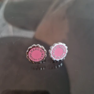 Rosa örhängen från Marc Jacobs - Säljer ett par söta örhängen från Marc Jacobs i silverfärg med rosa rund mitt och blomformad kant. Märkets logga står runt kanten. Perfekta för att ge en färgklick till din stil!