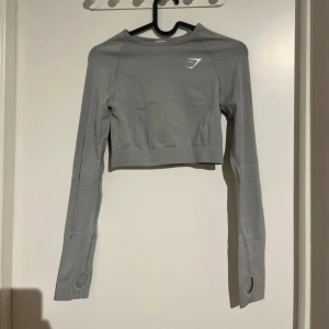 Grå croppad träningströja från Gymshark - Säljer en ljusgrå croppad långärmad träningströja från Gymshark. Tröjan har tumhål i ärmsluten och är tillverkad i ett stretchigt syntetmaterial som andas. Perfekt för gym eller annan träning.  Gymshark-logga på bröstet och rygg. 
