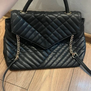 Väska  - Snygg svart handväska med quiltat mönster och guldfärgade kedjor på axelremmen. Väskan har ett elegant lock och två handtag, perfekt för att lyfta vilken outfit som helst. Materialet är skinnimitation och designen är stilren och modern.