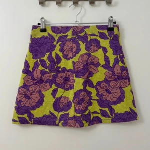 Blommig kjol från Zara - Säljer en kort kjol från Zara med stort blommigt mönster i lila och rosa på. Kjolen har en rak passform och dekorativa fickor bak. 