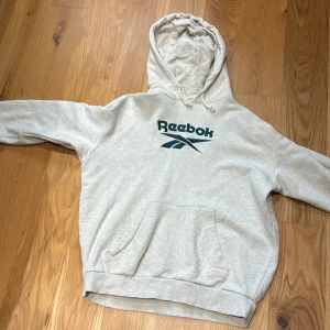 Ljusgrå hoodie från Reebok - Säljer en ljusgrå hoodie från Reebok med mörkgrön logga på bröstet. Tröjan har huva med  magficka framtill. Perfekt för en avslappnad stil. köpt för 500+ men säljer för 150