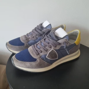 Philippe Model sneakers i blått och grått - Säljer ett par snygga philippe modells i blått och grått med gula detaljer på hälen. Skorna har snörning och en platt sula. Perfekta för dig som gillar en grisch och stilren look. Och dom är i nyskick. Ingen box eller og tillkommer tyvärr 