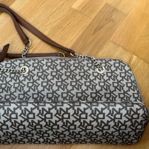 Brun och beige axelväska från DKNY - Säljer en snygg axelväska från DKNY med brunt och beige mönster och kedjedetaljer. Väskan har flera fack inuti och dragkedja upptill. Perfekt för dig som vill ha en stilren och praktisk accessoar.