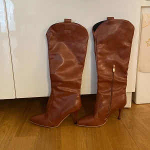 Bruna höga stövlar med klack - Snygga bruna höga stövlar i skinn med spetsig tå och smal klack. Stövlarna har dragkedja på  en sida och en stilren design som passar perfekt till höst och vinterlooken.
