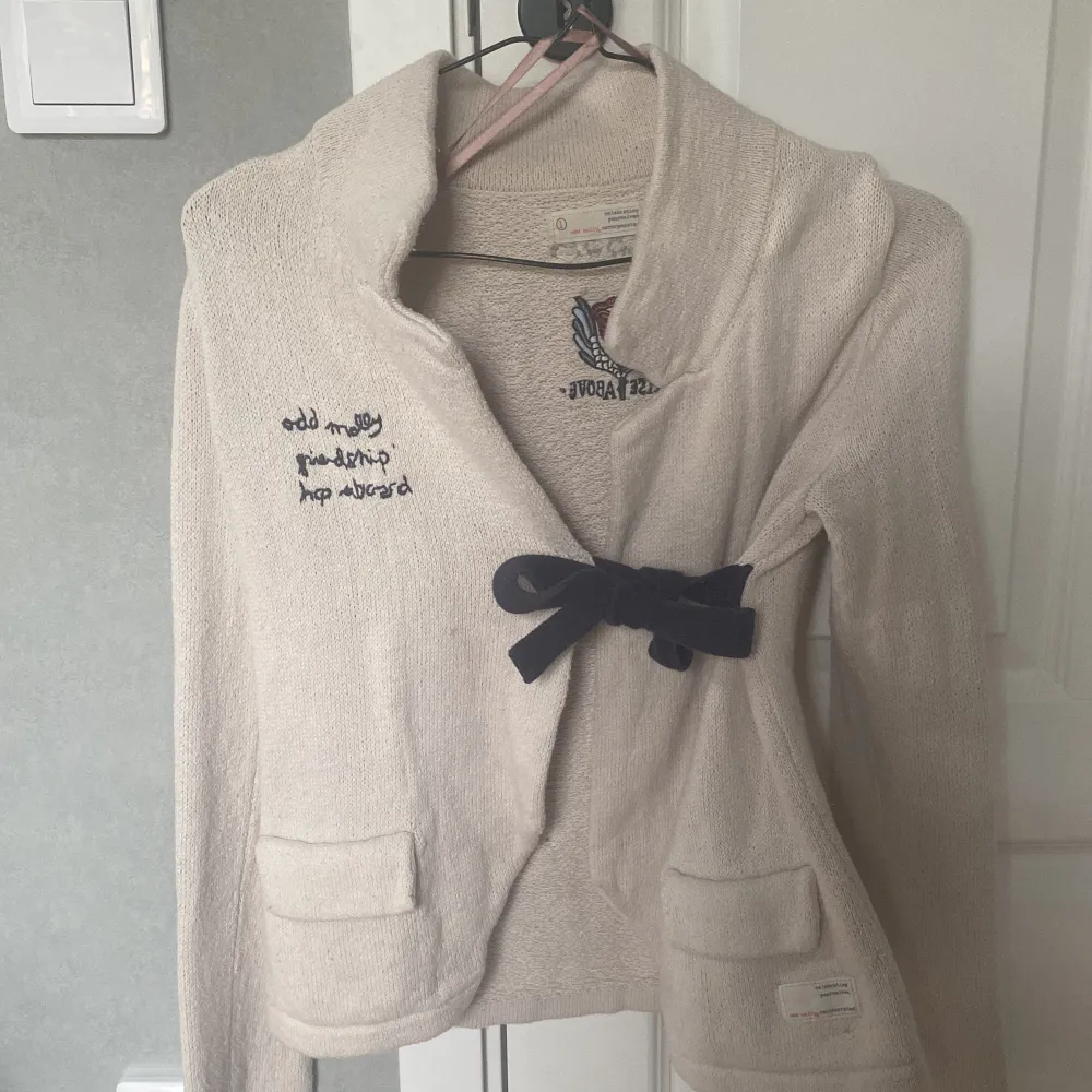 Säljer en beige stickad kofta från Odd Molly med broderad ros och texten 'Rise Above' på bröstet. Koftan har lång ärm, två fickor fram och knytdetalj i midjan. Perfekt för lager-på-lager och snygg till jeans eller kjol.. Neuletakit & Villapaidat.