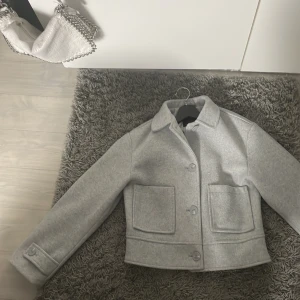 Ljusgrå kappa  - Säljer en stilren ljusgrå kappa i storlek XS. Bara använd nån eller några fåtal gånger. Kappan har klassisk krage, stora framfickor och knäppning med knappar framtill.