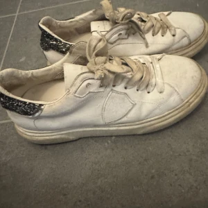 Vita sneakers från Philippe Model  - Säljer ett par vita sneakers från Philippe Model med svart glittrig detalj på hälen. Skorna har snörning och en klassisk rund tå. Perfekta för dig som gillar en stilren look med en touch av glitter.