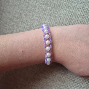 Lila armband med pärlor - Handgjort armband med vita pärlor och lila pärlor runtom. Pärlorna är jämnt placerade i mitten och omges av lila pärlor som ger ett unikt och stilrent utseende. Perfekt accessoar för att addera lite färg och elegans till din outfit.