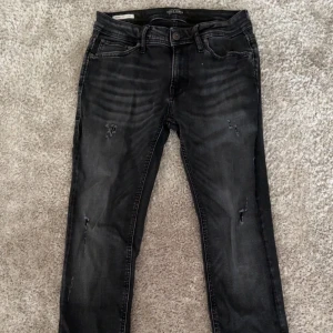 Svarta jeansbyxor med slitningar - Säljer ett par svarta jeansbyxor med diskreta slitningar på benen. Byxorna har klassisk femficksdesign och knappgylf. Snygg tvättad look som passar till många stilar. Storlek W29 L32, modell skinny fit men sitter slim