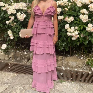 Ljusrosa långklänning med volanger - Romantisk ljusrosa långklänning med flera lager volanger och smala axelband. Djup v-ringning och spetsdetalj i midjan. Perfekt för somriga tillfällen och studentfirande.