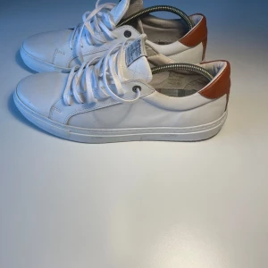 Vita sneakers från Barbour med små bruna detaljer - Snygga vita sneakers från Barbour med bruna detaljer på hälen. Skorna har rund tå, platt sula och klassisk snörning. Perfekta för en stilren och avslappnad look. Märkeslogga på plösen och insidan. 