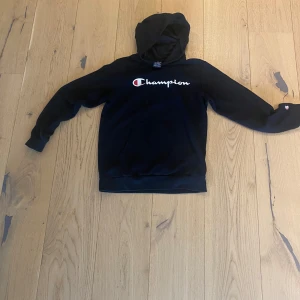 Svart hoodie från Champion - Säljer en svart hoodie från Champion med klassisk logga broderad på bröstet. Tröjan har huva och magficka, samt långa ärmar. Perfekt för en avslappnad stil.