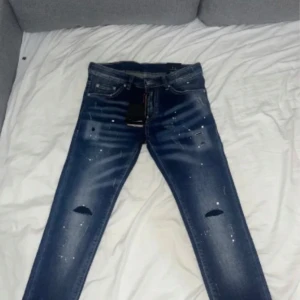 Blå slitna jeansbyxor från Dsquared2 - Säljer ett par blå jeansbyxor från Dsquared2 med slitningar, hål på knäna och vita färgstänk. Klassisk femficksmodell med normal passform och snygga detaljer. Märkeslapp bak i midjan. Perfekta för en trendig streetstil.