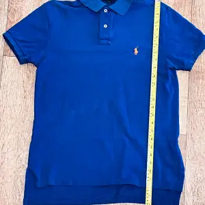 Säljer en klassisk blå piké t-shirt från Ralph Lauren med orange broderad logga på bröstet. T-shirten har krage, två knappar vid halsen och är kortärmad. Perfekt för en stilren och sportig look. Snabb leverans 