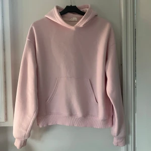 Ljusrosa hoodie - Säljer en ljusrosa hoodie med huva och magficka. Tröjan har en avslappnad passform och ribbade muddar vid ärmslut och nederkant. Perfekt för en chill och bekväm stil.