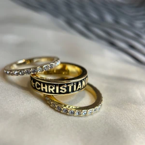 Guldfärgade ringar från Dior med stenar - Säljer tre snygga guldfärgade ringar från Dior. En ring har texten 'CHRISTIAN DIOR' i svart och guld, de andra två är dekorerade med gnistrande stenar runt hela ringen. Perfekta för att ge din look lite extra lyx. Size M, FR52