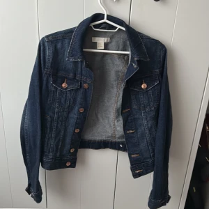 Mörkblå jeansjacka från H&M - Säljer en klassisk mörkblå jeansjacka från H&M i storlek XS. Jackan har kopparfärgade knappar, två bröstfickor med lock och knapp samt snygga söm-detaljer. Perfekt att slänga över en hoodie eller t-shirt.