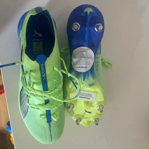 Puma Ultra fotbollsskor i neonfärg - Säljer ett par Puma Ultra fotbollsskor i neonljusgrön och blå färg med svarta detaljer. Skorna har snörning och metall-dobbar för bästa grepp på planen. Perfekta för dig som vill sticka ut på fotbollsplanen!