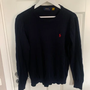 Svart stickad tröja från Polo Ralph Lauren - Klassisk svart stickad tröja från Polo Ralph Lauren med rund halsringning och röd broderad logga på bröstet. Långärmad modell med ribbade muddar vid ärmslut och nederkant. Perfekt för lager-på-lager-stil.