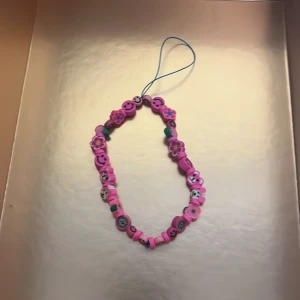 Rosa pärlarmband med smileys och blommor - Färgglatt armband med rosa och lila pärlor, dekorerat med smileys och blommor. Armbandet har elastisk tråd och en blå ögla för enkel användning. Perfekt accessoar för att liva upp din stil.