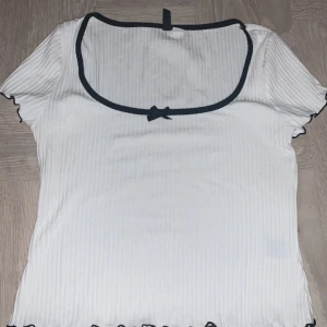 Vit ribbad topp med svart kant och rosett - Söt vit ribbad topp med svart kant runt den fyrkantiga ringningen och en liten svart rosett framtill. Toppen har korta ärmar och vågig kant nertill. Perfekt för en trendig och stilren look.