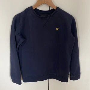 Mörkblå sweatshirt från Lyle & Scott - Klassisk mörkblå sweatshirt från Lyle & Scott med rund halsringning och broderad gul logga på bröstet. Tröjan är varsamt använd och inga defekter. Storleken är 12/13 år vilket motsvarar ungefär 164. Tröjan har långa ärmar och ribbade muddar vid ärmslut och nederkant. Perfekt för en avslappnad stil men som även är stilren.