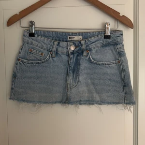 Ljusblå jeanskjol från Gina Tricot - Säljer en ljusblå jeanskjol från Gina Tricot i kort modell med klassisk femficksdesign och rå, fransig nederkant. Kjolen har bälteshällor och knappstängning framtill. Längd 22/26 cm
