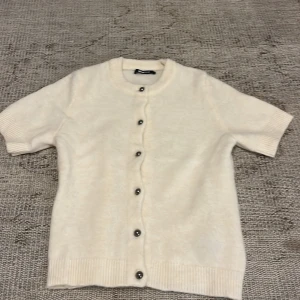 Vit kofta från Gina Tricot - Säljer en söt vit kofta från Gina Tricot i storlek XS. Koftan har korta ärmar, rund halsringning och knappar framtill. Perfekt att styla över en topp eller klänning för en fräsch look.
