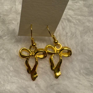 Guldiga örhängen med rosettform - Säljer ett par snygga örhängen i guldmetall med en elegant rosettform. Perfekta för att ge din outfit en söt och trendig touch. Hängande modell med krokfäste. Krokarna är i sterling silver 