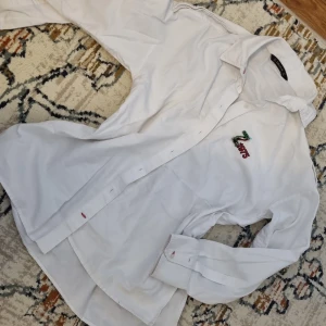 Vit skjorta från Zara Basic med broderad detalj - Säljer en klassisk vit skjorta från Zara Basic i bomull. Skjortan har lång ärm, knappar framtill och en broderad detalj med texten 'Zara' i färg på bröstet. Perfekt för en stilren och enkel look.