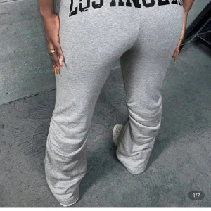 Grå mjukisbyxor med textdetalj - Säljer ett par grå mjukisbyxor med svart textdetalj bak på midjan. Byxorna har normal passform, hög midja och utsvängda ben med rynkade detaljer längs sidorna. Perfekta för en avslappnad och trendig look.