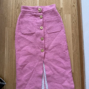 Rosa långkjol med guldknappar - Säljer en snygg rosa långkjol med dekorativa guldknappar framtill och två stora fickor. Kjolen har en rak passform och en slits framtill för extra rörelsefrihet. Perfekt för dig som vill sticka ut med färg!