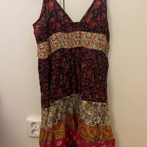 Flerfärgad långklänning med blommigt mönster - Säljer en bohemisk långklänning med smala axelband och flera olika blommiga mönster i svart, rosa, orange och beige. Klänningen har v-ringning och justerbara axelband. Perfekt för sommar😍 