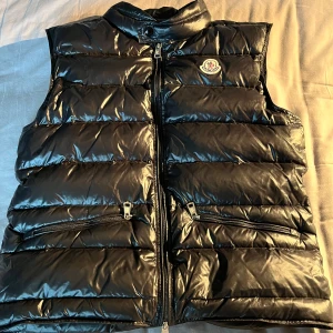 Moncler gui - Säljer en svart, glansig dunväst från Moncler med hög krage och dragkedja framtill. Västen har två snedställda fickor med dragkedja och Moncler-logga på bröstet. Perfekt för lager-på-lager och stilren look. Storlek 1, passar från folk som e 150 till 170cm