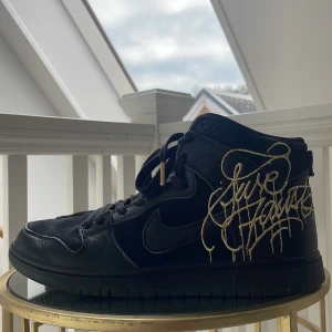 Nike SB Dunk High x FAUST Black Metallic Gold Graffiti Street Art Shoes  - Säljer ett par svarta Nike Dunk High sneakers med unika guldfärgade grafitti-detaljer på sidan och guldfärgad swoosh. Skorna har snörning och är tillverkade i skinn. Perfekta för dig som vill sticka ut med en exklusiv look.