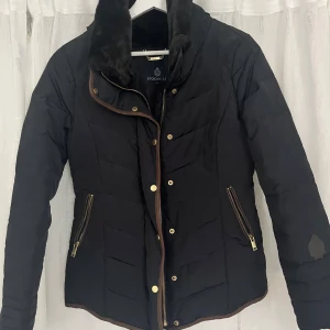 Svart quiltad jacka med fuskpäls från Stockh LM - Snygg svart quiltad jacka från Stockh LM med mjuk fuskpäls vid kragen och guldfärgade dragkedjor. Jackan har långa ärmar, bruna detaljer längs kanterna och två fickor med dragkedja framtill. Perfekt för kyliga dagar.