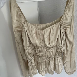 Beige långärmad blus med volang - Säljer en beige långärmad blus med v-ringning och volangdetaljer. Blusen har en lätt glansig yta och smockade partier som ger en fin passform. Perfekt att styla med jeans eller kjol.