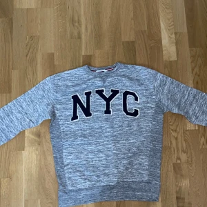Grå sweatshirt med NYC-tryck - Säljer en gråmelerad sweatshirt med stort svart NYC-tryck på bröstet. Tröjan har rund halsringning och långa ärmar. Perfekt för en avslappnad stil.