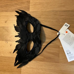 Svart maskeradmask med spets och fjäderdetaljer från H&M - Snygg svart maskeradmask med spets och dekorativa fjäderliknande detaljer runt ögonen. Masken har ett elastiskt band för att sitta bekvämt på plats. Perfekt för maskerad eller fest.
