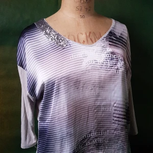 Randig topp med paljettdetalj - Säljer en snygg topp med grå och vita ränder samt texttryck. U-ringad hals med silvriga paljetter på ena sidan. Trekvartsärm och lätt glansigt material. Perfekt för dig som vill sticka ut lite extra.