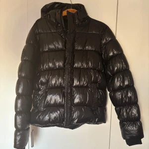 Svart pufferjacka från Superdry XP-D - Säljer en svart pufferjacka från Superdry XP-D med huva och dragkedja. Jackan har en glansig finish och vadderad design som håller värmen. Perfekt för kyliga dagar och har en snygg logga på ärmen och ryggen. Jackan är sparsamt använd under endast en vinter. 