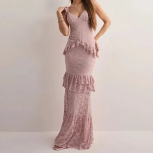 Ljusrosa långklänning med volang och spets - Romantisk ljusrosa långklänning med smala axelband, v-ringning och volangdetaljer. Klänningen är figurnära och har spetsmaterial samt öppen rygg med snörning. Perfekt för speciella tillfällen.