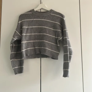Grå randig stickad tröja från Zara - Säljer en grå stickad tröja från Zara med vita horisontella ränder. Tröjan är croppad med lång ärm och har en ribbad struktur. Perfekt för dig som gillar en avslappnad och trendig stil. Ett litet hål finns men inget som märks mycket.