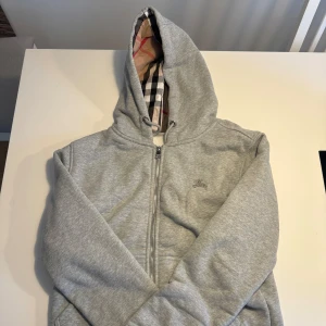 Grå Burberry zip hoodie - Grå Burberry zip hoodie med mönstret på luvan samt inuti. Denna är i Storlek S, skick 10/10. Vårt pris: 999kr. Hör av er vid minsta fråga!