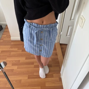 Blåvit randig kjol med knytband och knappar - Säljer en blå kjol med vita ränder och knappar framtill. Kjolen har ett knytband i midjan och är i kort modell. Perfekt för en avslappnad och somrig look. Är i storlek 146-152 men passar som en xs. 