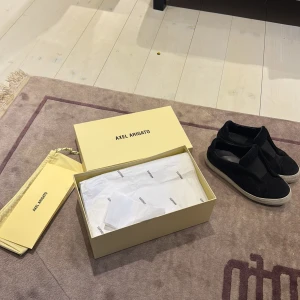 Axel Arigato 360 laceless - Säljer ett par svarta slip-on sneakers från Axel Arigato med vit platt sula. Skorna har en minimalistisk design utan snörning och är tillverkade i mocka. Perfekta för en clean och stilren look.